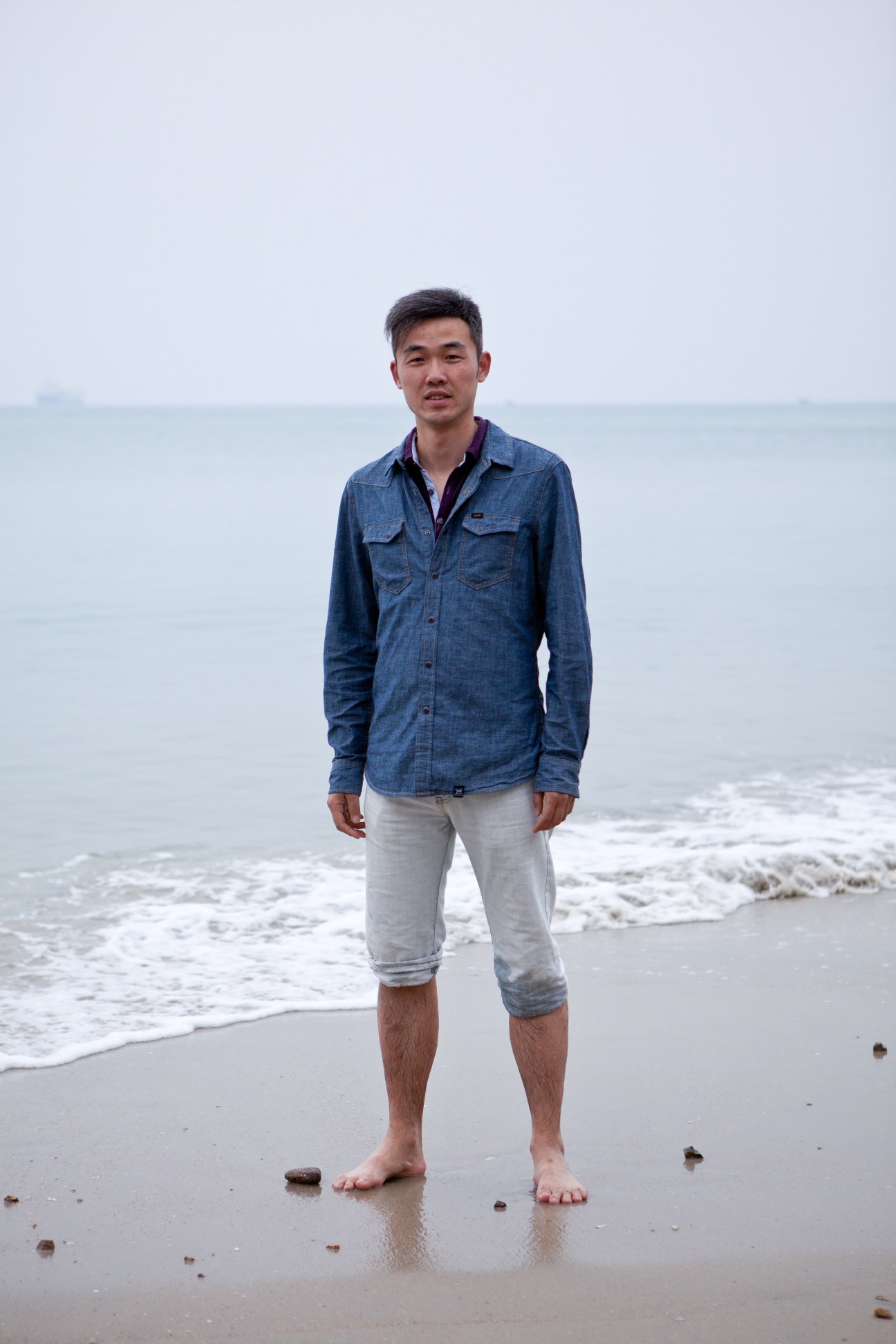 Man standing barefoot on beach in Sanya wearing denim shirt and light pants.