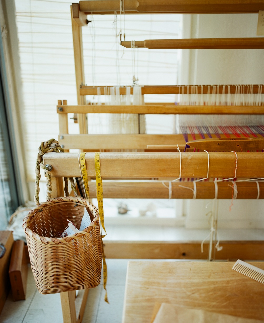 Wicker basket hanging from a wooden loom with yarns and weaving materials visible.