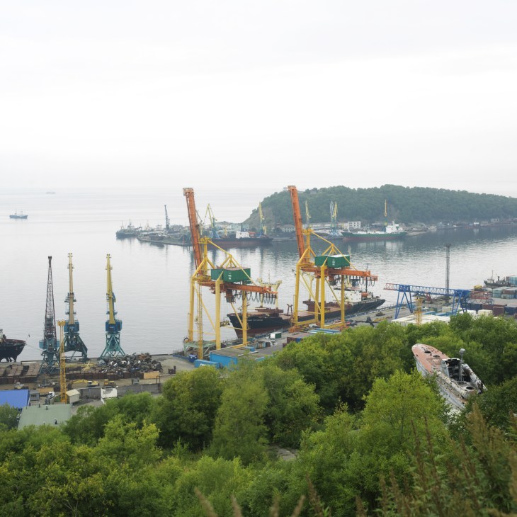 Industrial docks at Petropavlovsk with yellow cranes, cargo ships, and forested hillside.