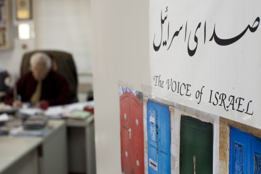 Sign reading 'The Voice of Israel' in Farsi and English on an office wall with a person working at a desk in the background.