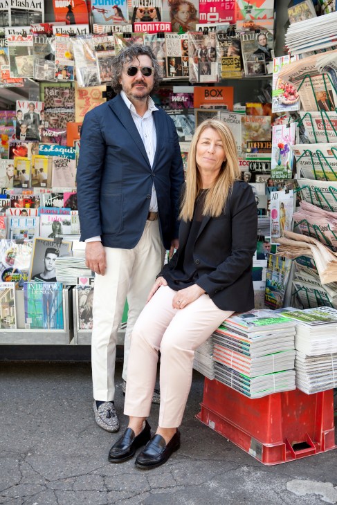 Two people stand in front of a magazine and newspaper kiosk display in Milan.