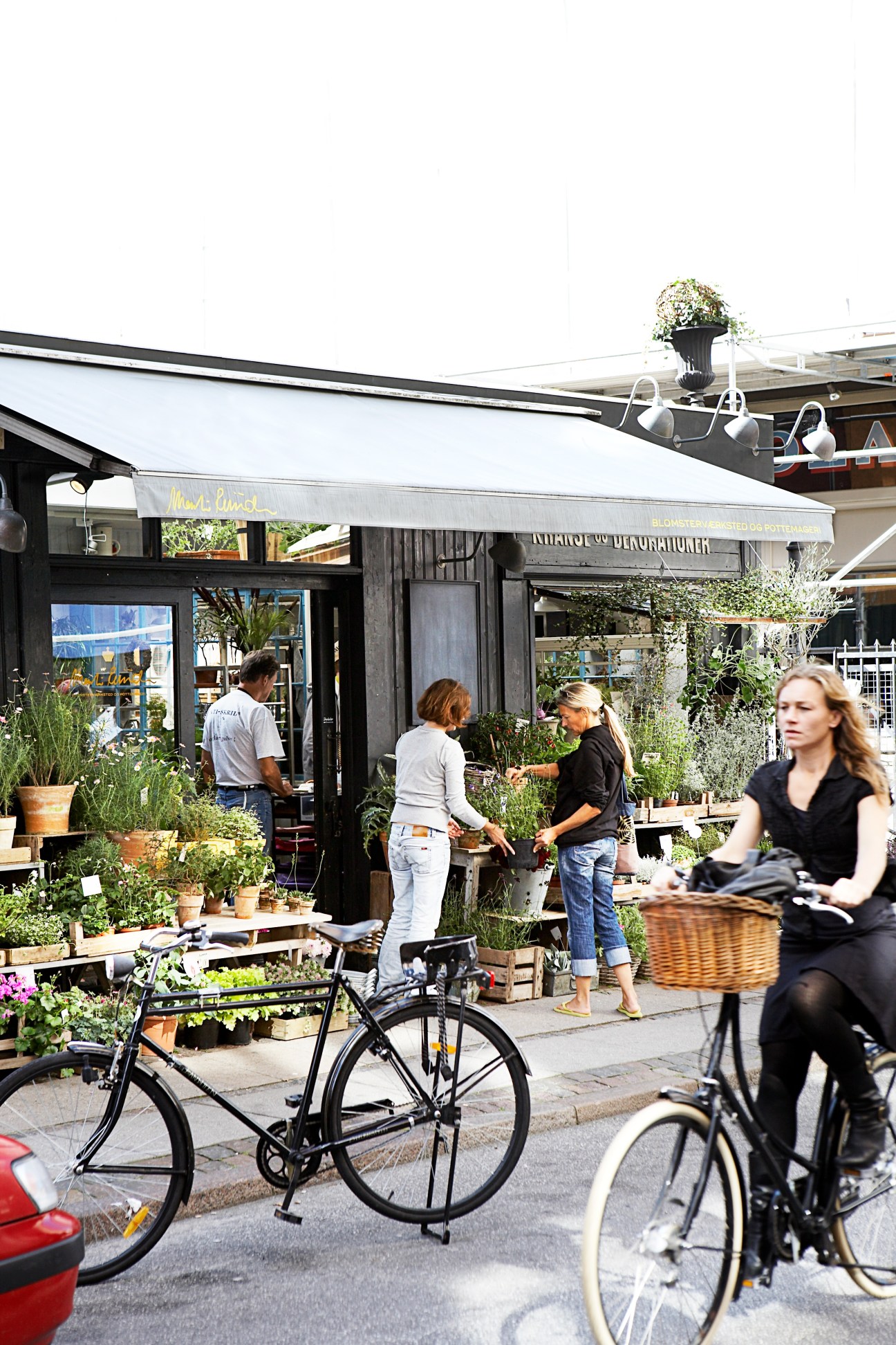 Street scene in Frederiksberg neighbourhood with cyclists, pedestrians, and a black storefront with white awning surrounded by potted plants.