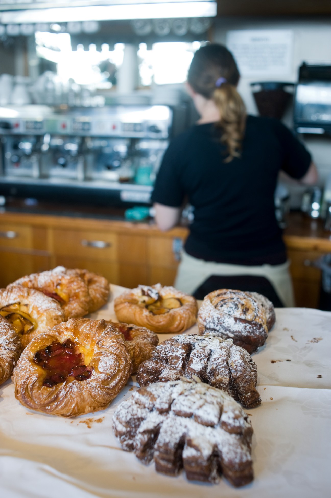 Assorted pastries including croissants and chocolate-dusted baked goods displayed in a café with barista working in background.