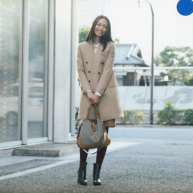 Woman wearing a beige Gucci coat with a patterned Gucci bag, standing in front of modern glass doors.