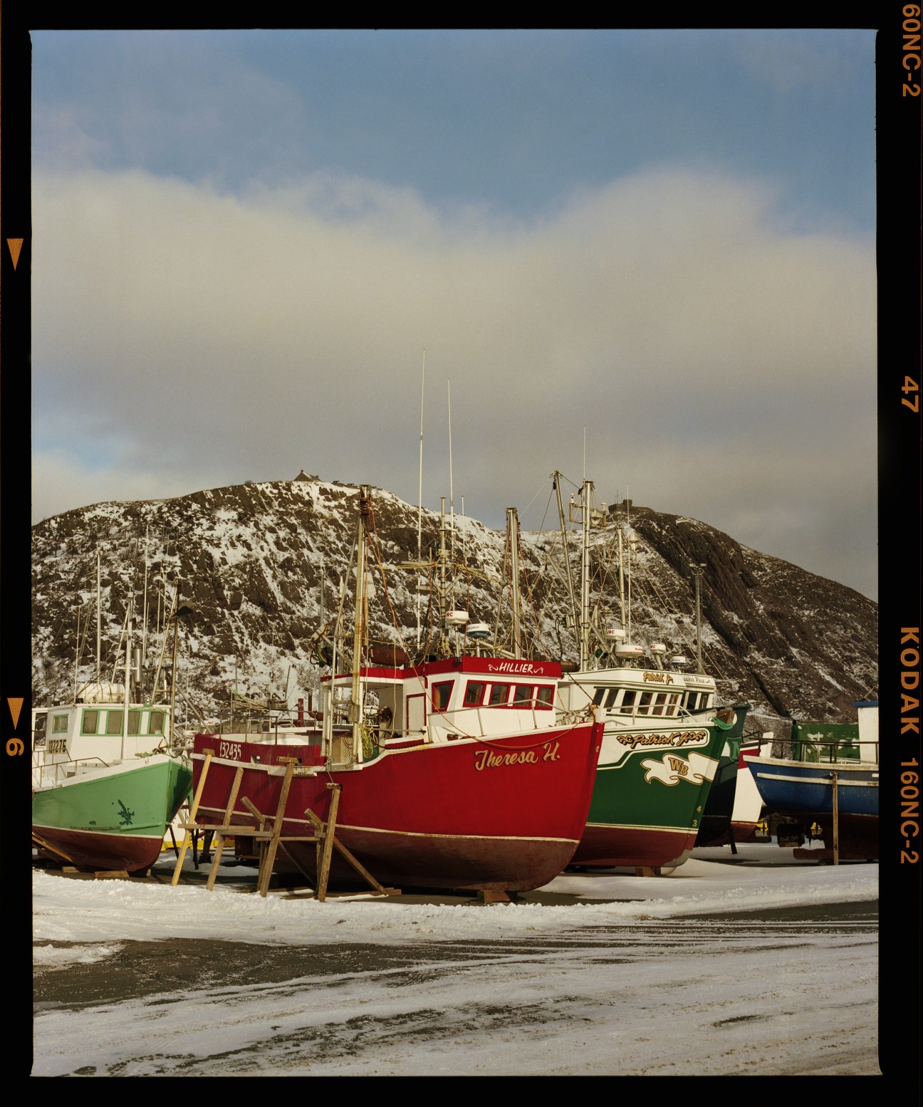 Colorful fishing boats moored on a beach with snow-covered rocky hills in the background under a cloudy sky.