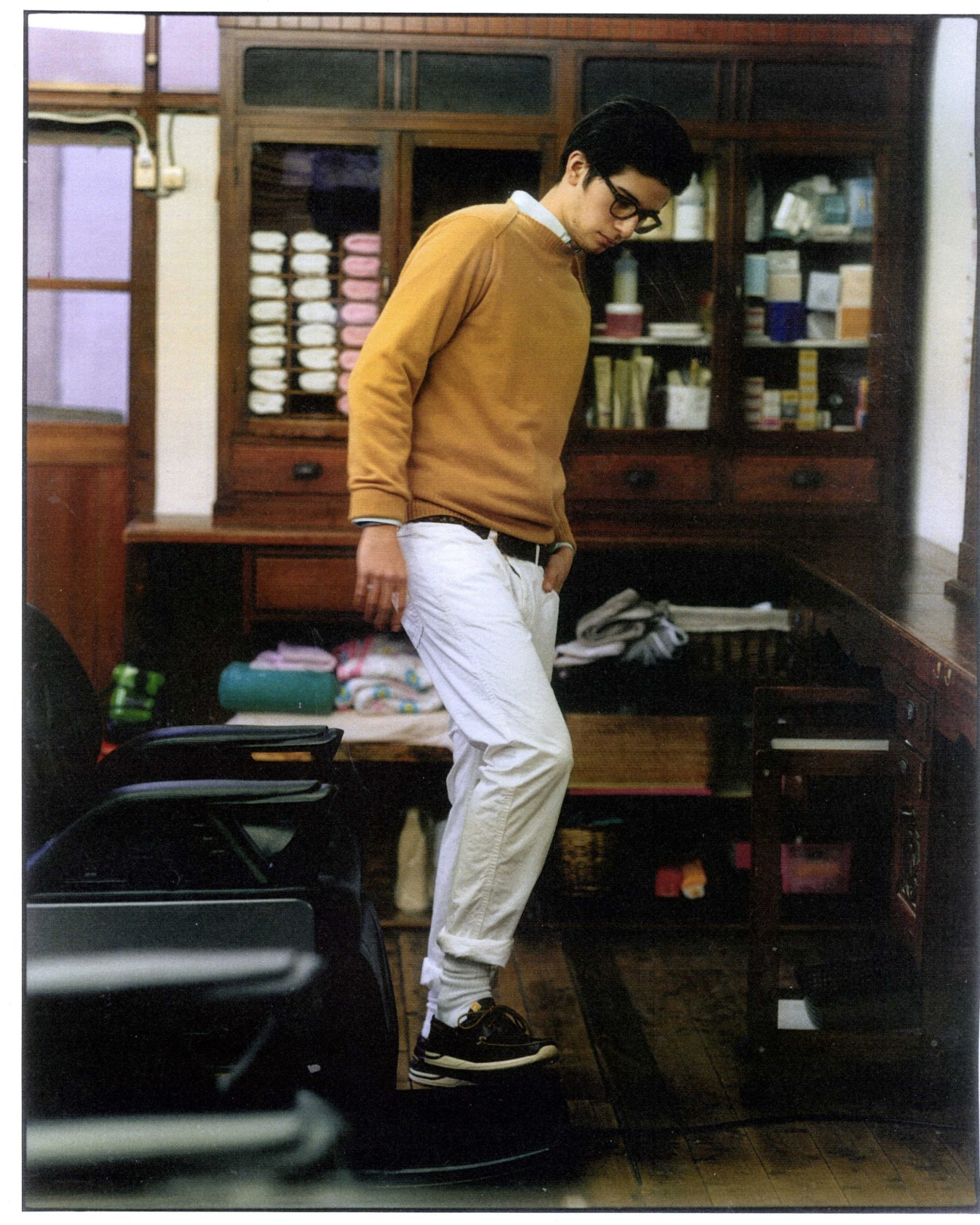 Man in yellow jumper and white trousers standing inside a traditional wooden-paneled barber shop with vintage cabinetry.