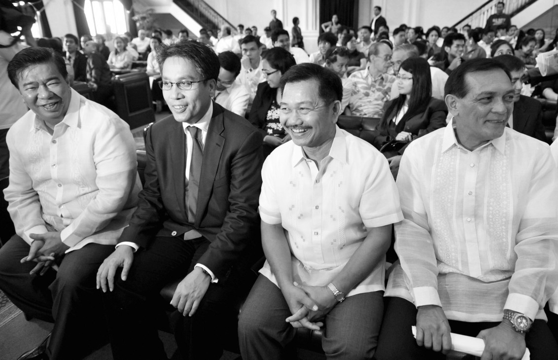 Manuel Roxas seated with three other men in formal white shirts at an indoor event with crowds visible in the background.