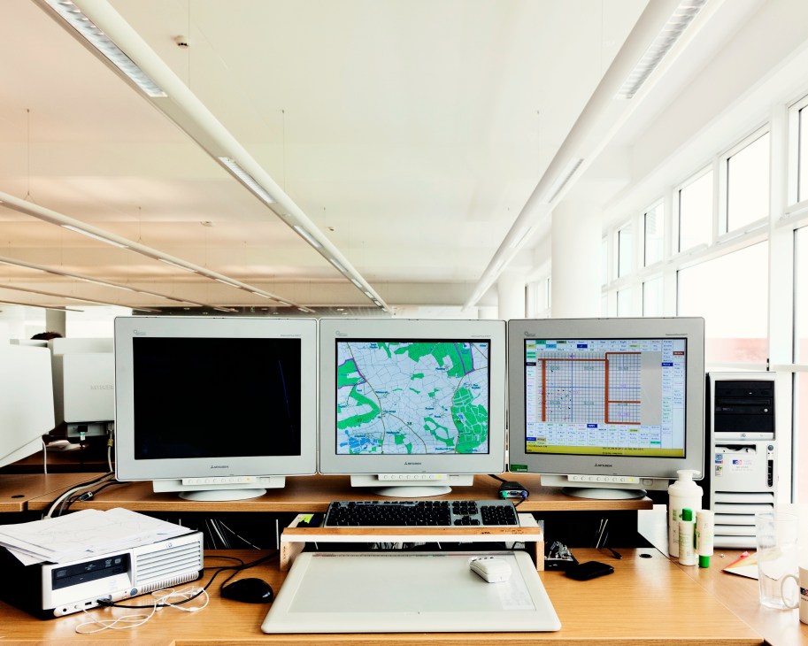 Three computer monitors displaying mapping software and aerial imagery on a desk in a bright office.