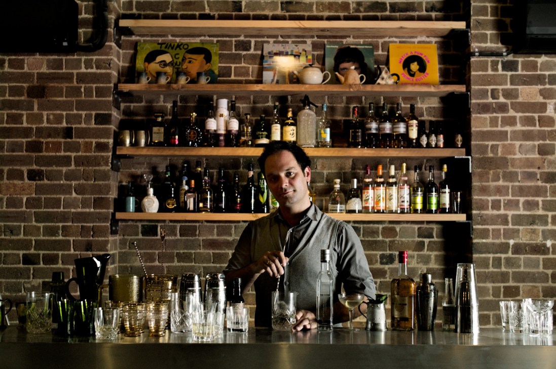 Bartender stands behind counter at Freda's bar with spirit bottles on shelves and colorful artwork displayed above.