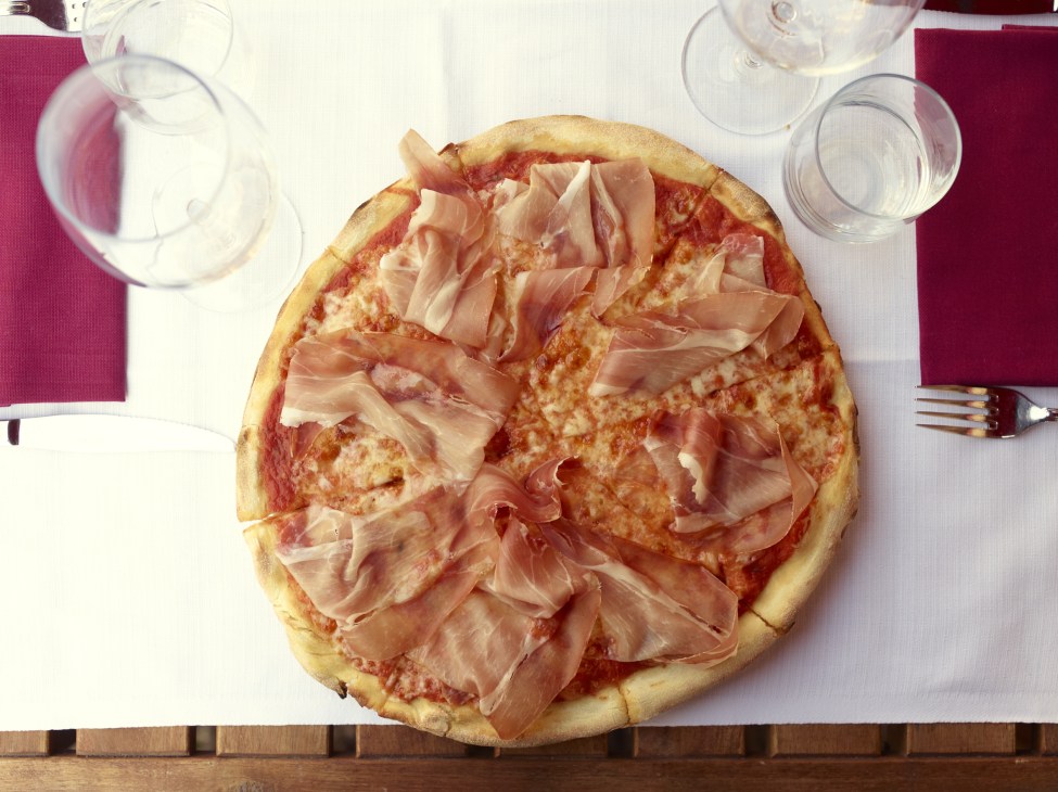 Margherita pizza with prosciutto di Norcia served on a white plate with water glasses and burgundy napkins.