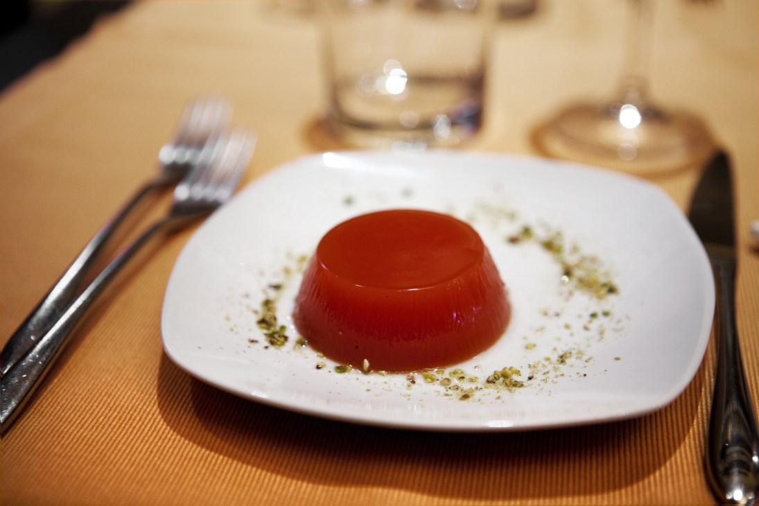 Red gelatin dessert garnished with chopped pistachios on a white plate with fork and knife.
