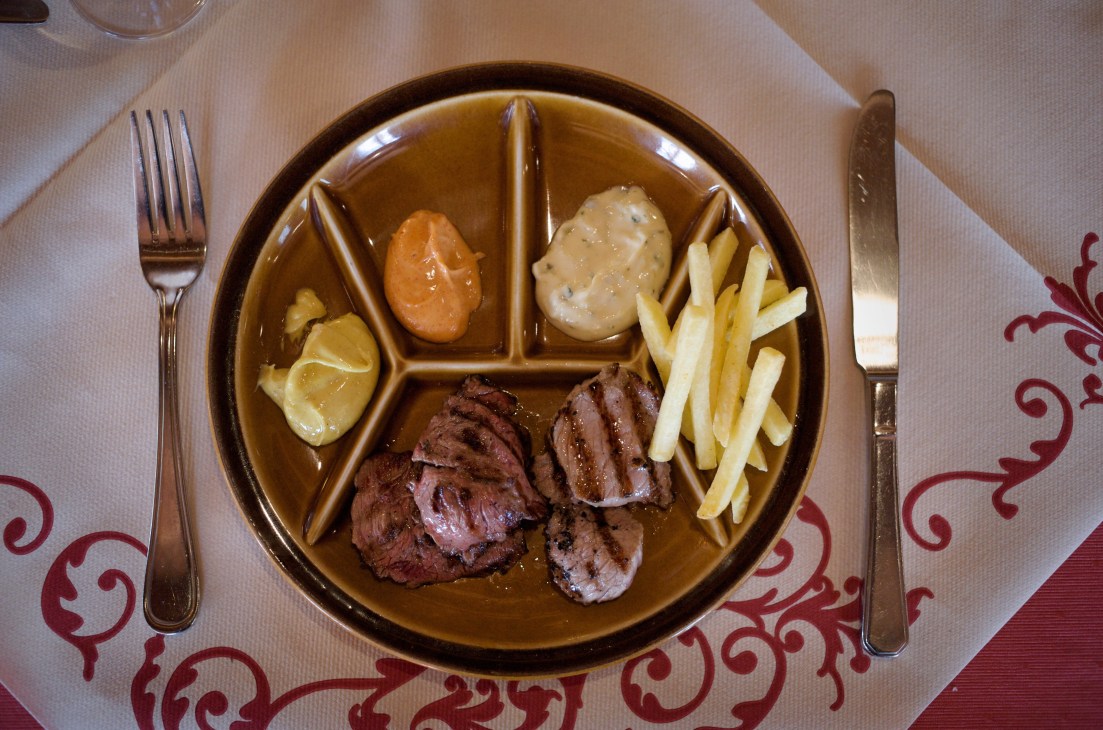 Cooked charbonnade beef, veal and bacon on a divided plate with fries and sauce compartments.