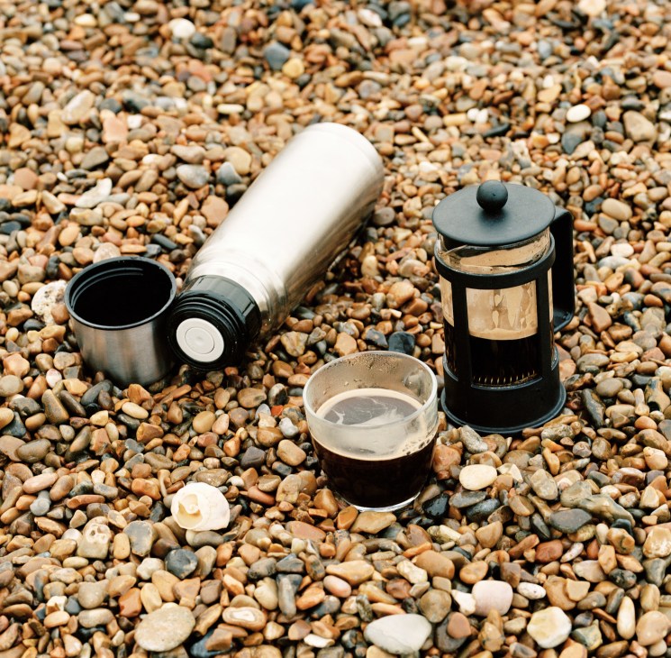 Coffee brewing setup on a pebble beach with a thermos flask, cafetière, metal cup and shells.