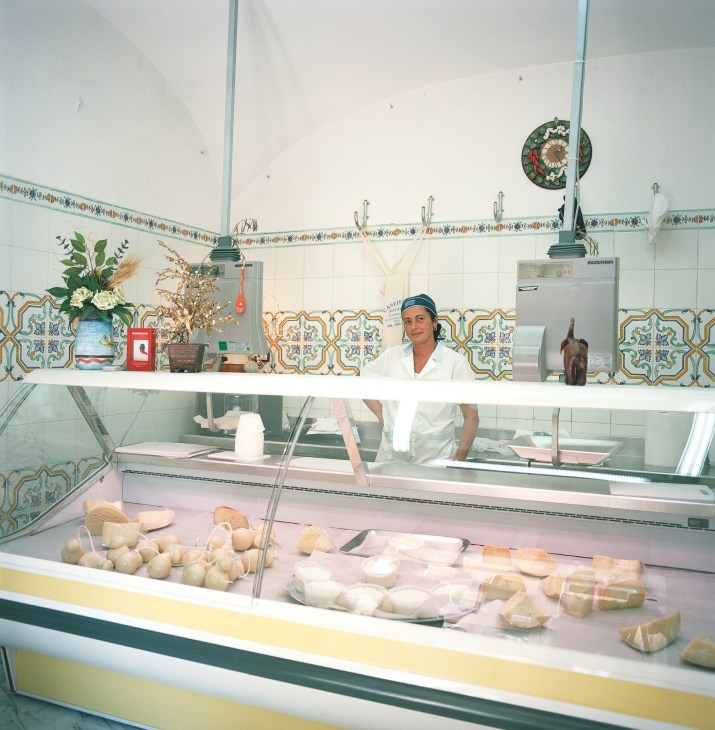 Woman behind counter at Caseificio dell'Artista cheese shop with display of mozzarella and decorative tiled walls.