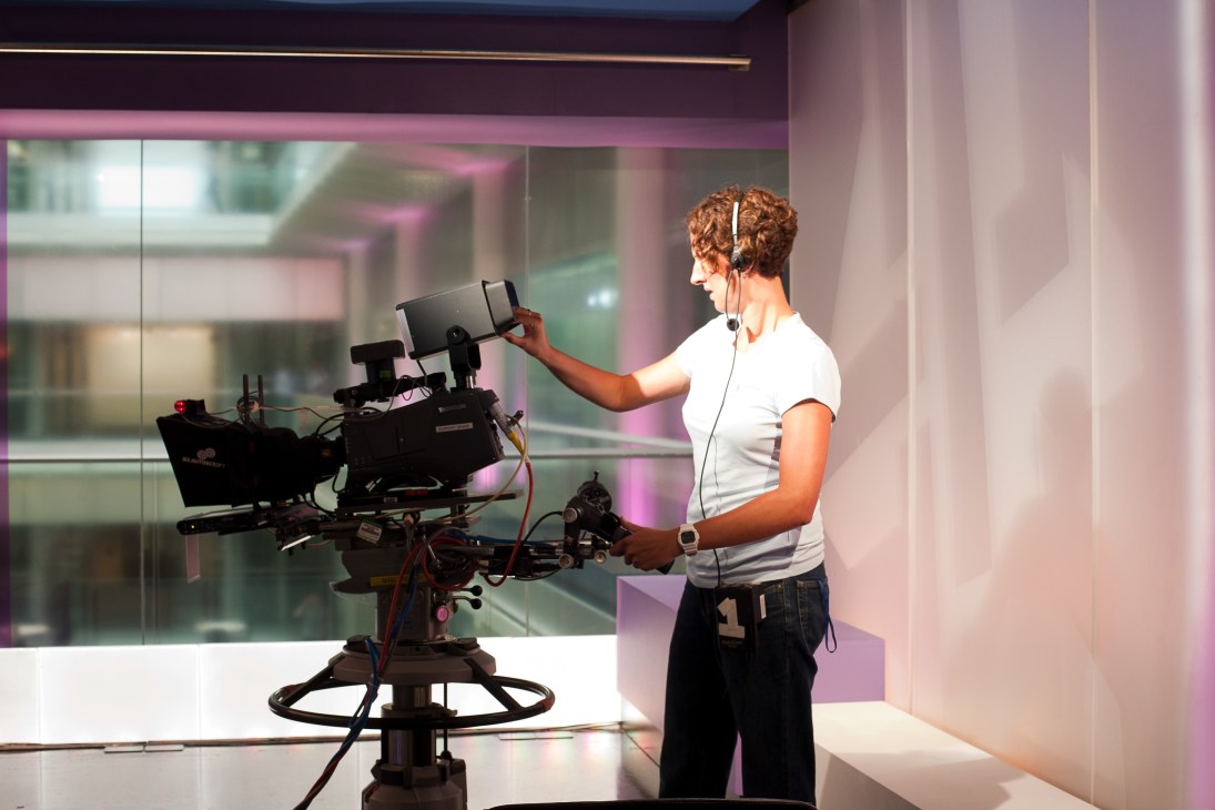 Female producer wearing headset adjusts camera equipment in a television studio with pink-purple ambient lighting.