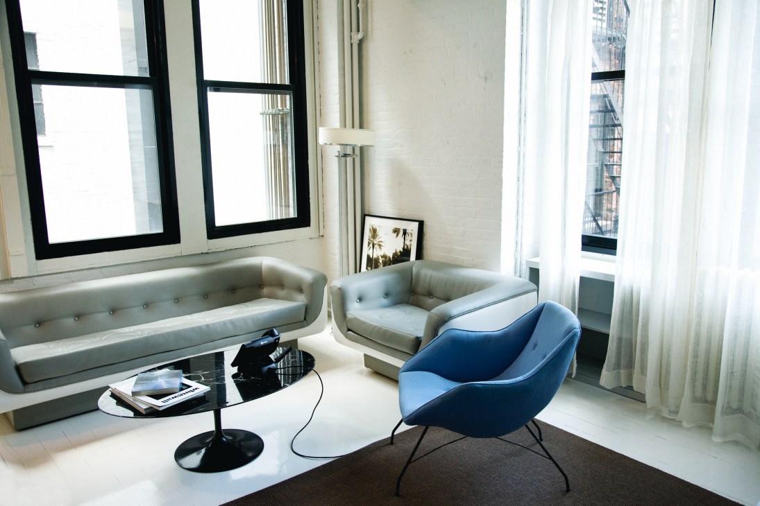 Modern seating area with gray upholstered sofa and chairs, blue accent chair, and black side table in bright office space with large windows.