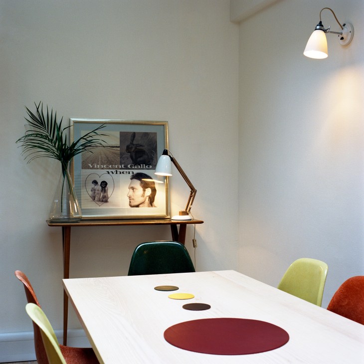 Meeting room with white table featuring colored circles, mid-century modern chairs, wall sconce, desk lamp, and framed artwork above wooden shelf.