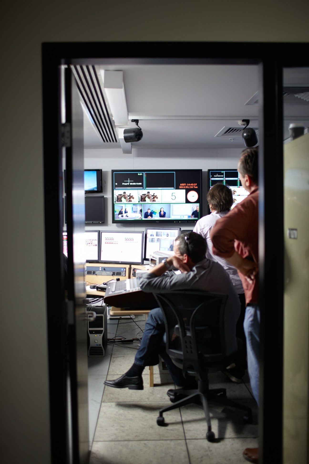 TV studio technicians work at a control room desk with multiple monitors displaying video feeds and graphics.