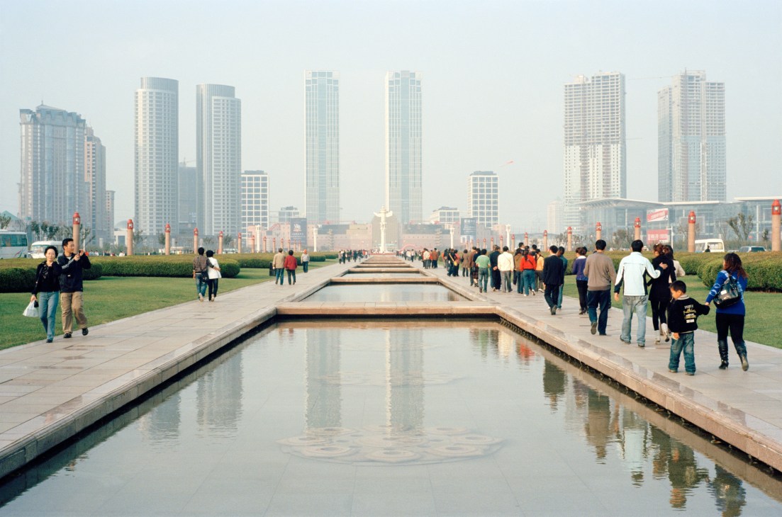 Xinghai Square in Dalian with a long reflecting pool, manicured lawns, and modern high-rise buildings under hazy skies.