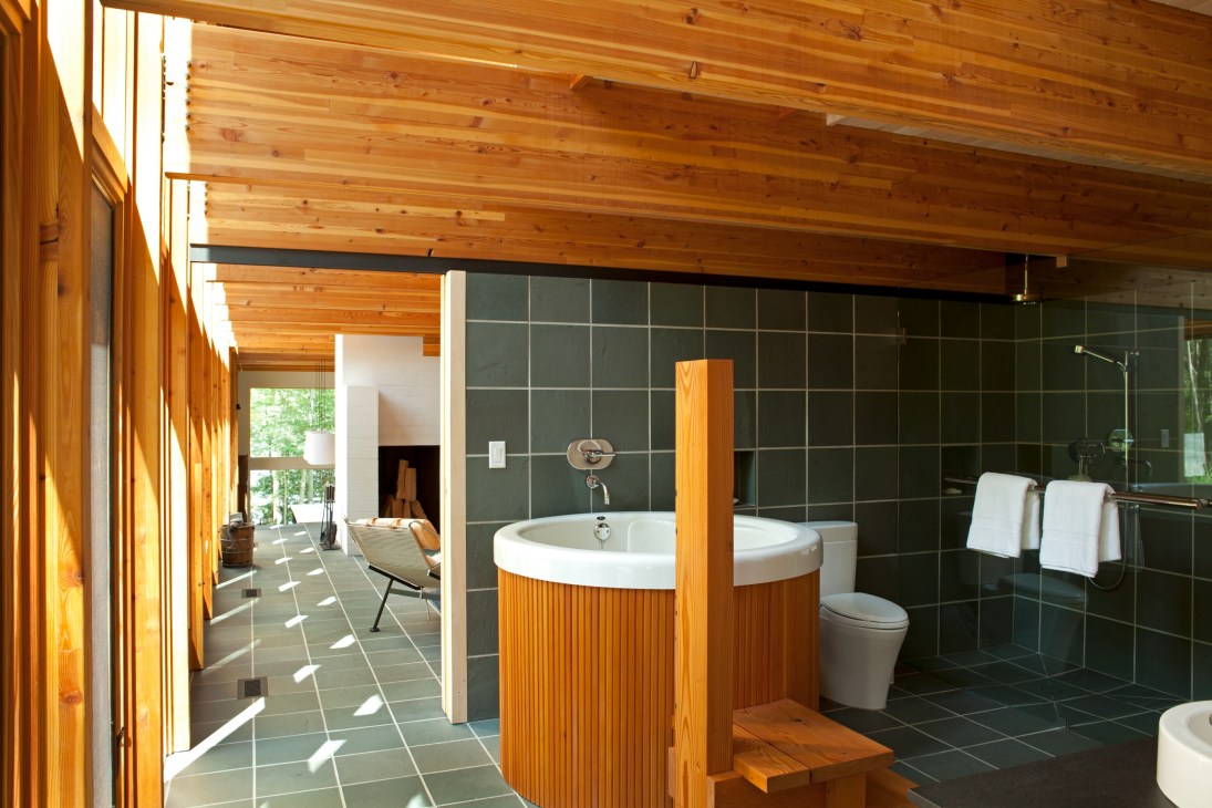 Modern master bathroom with wooden beams, round soaking tub, dark tile walls, and large windows overlooking forest.