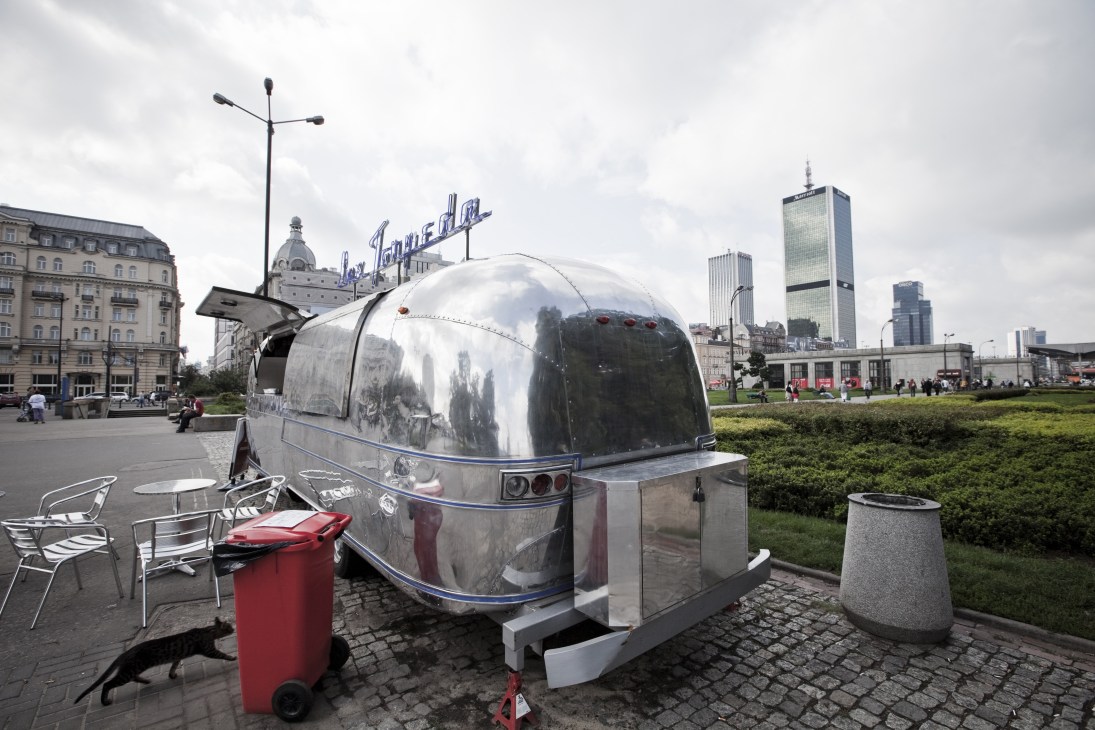 A vintage silver diner van with red accents parked in a Warsaw plaza, with modern skyscrapers visible in the background.