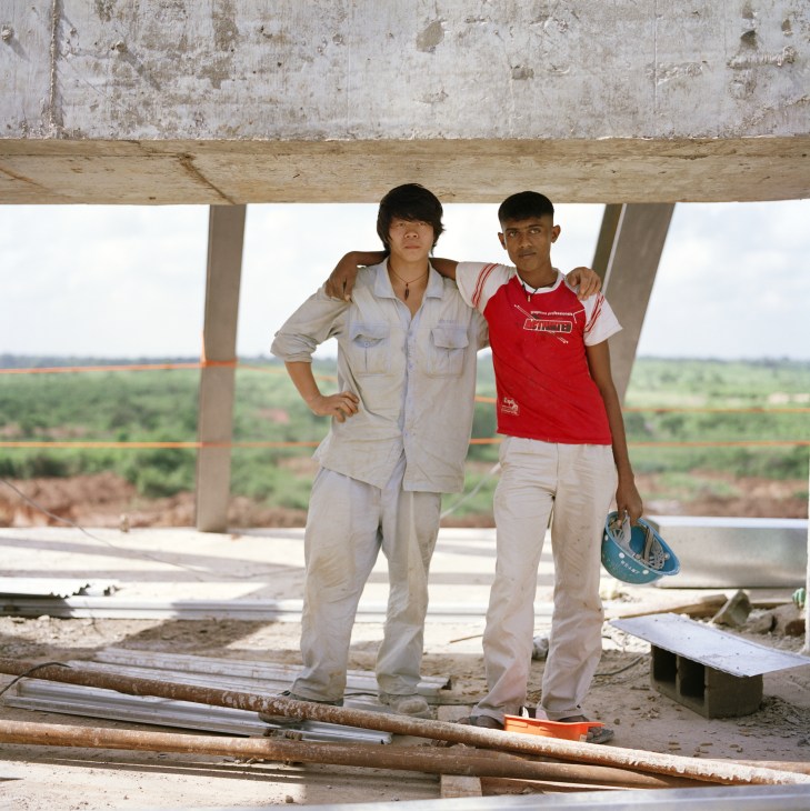 Two workers stand together under a wooden beam at a construction site with green fields visible in the background.