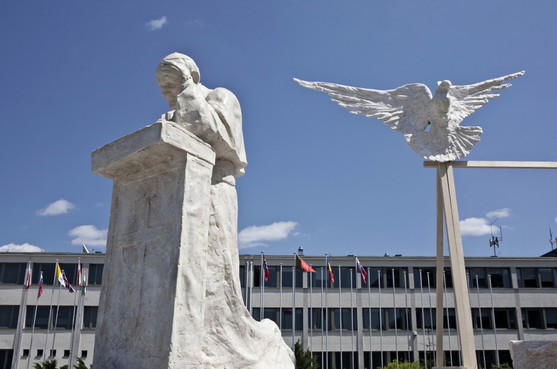 White marble statue of Pope John Paul II holding a cross, with a dove sculpture to the right against a blue sky.