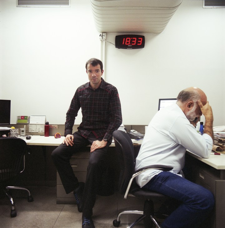Two men in an office setting; one sits on desk edge in plaid shirt, the other sits at desk in white shirt with hand to face.