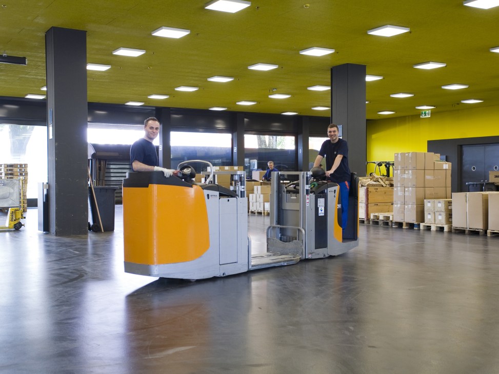Workers operate robotic forklifts in V-Zug's bright yellow-ceilinged logistics storage facility.
