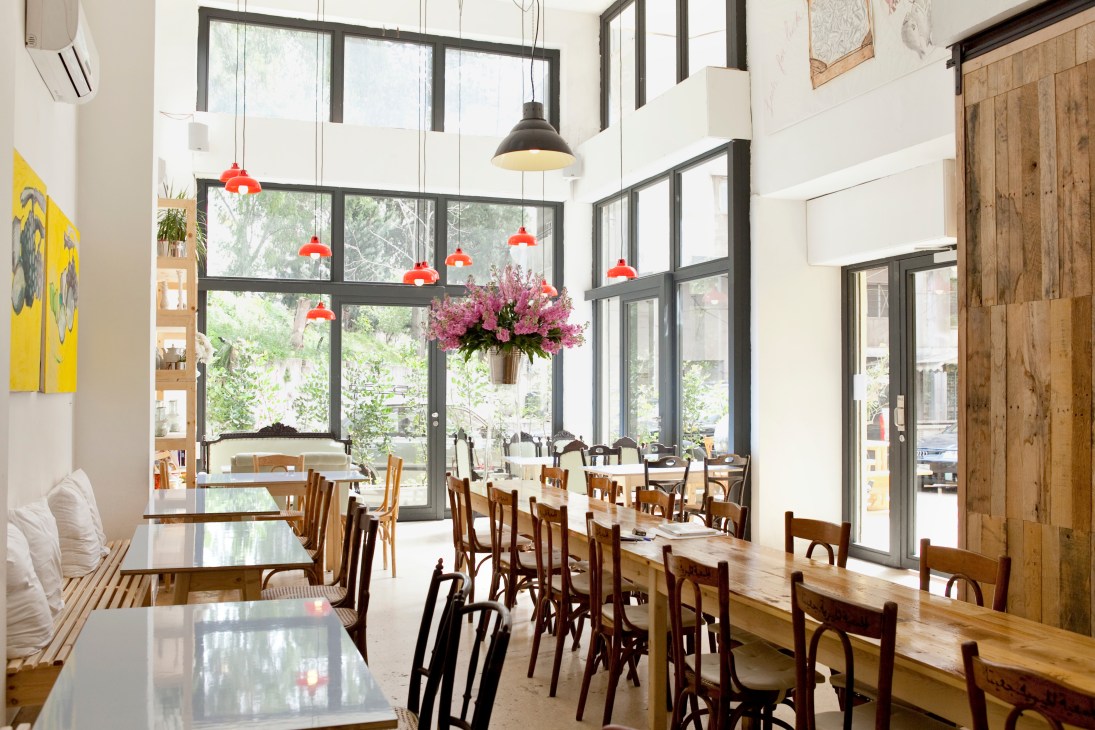 Bright restaurant interior with wooden tables, mid-century chairs, hanging red pendant lights, and large windows overlooking greenery.