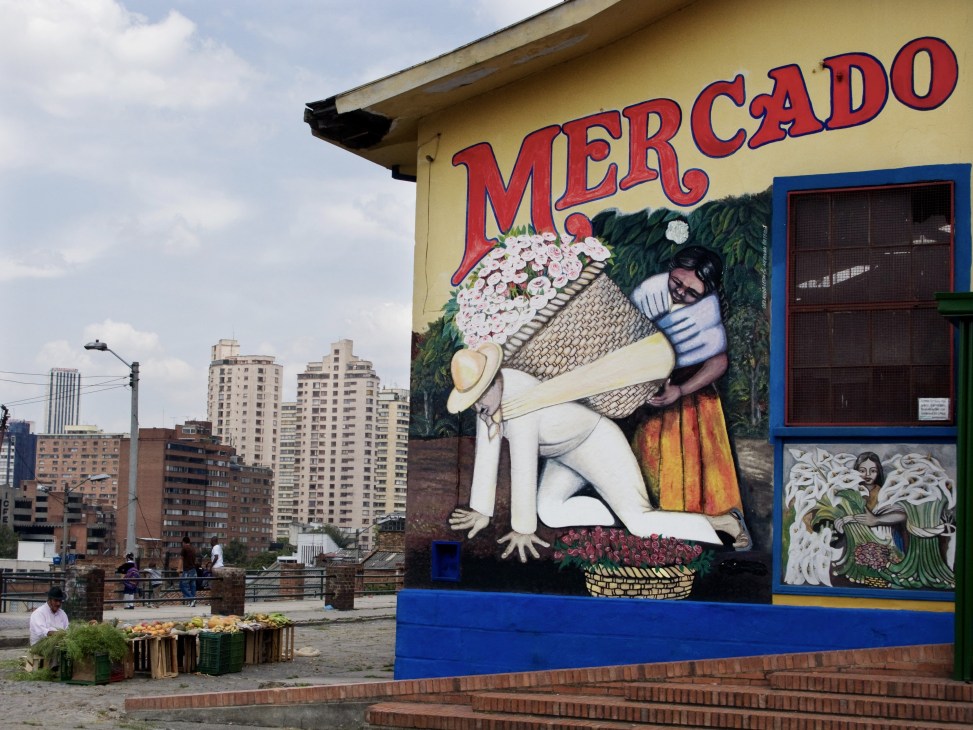 Colorful mural at Plaza de Mercado featuring a figure holding flowers and produce, with Bogotá skyline behind.