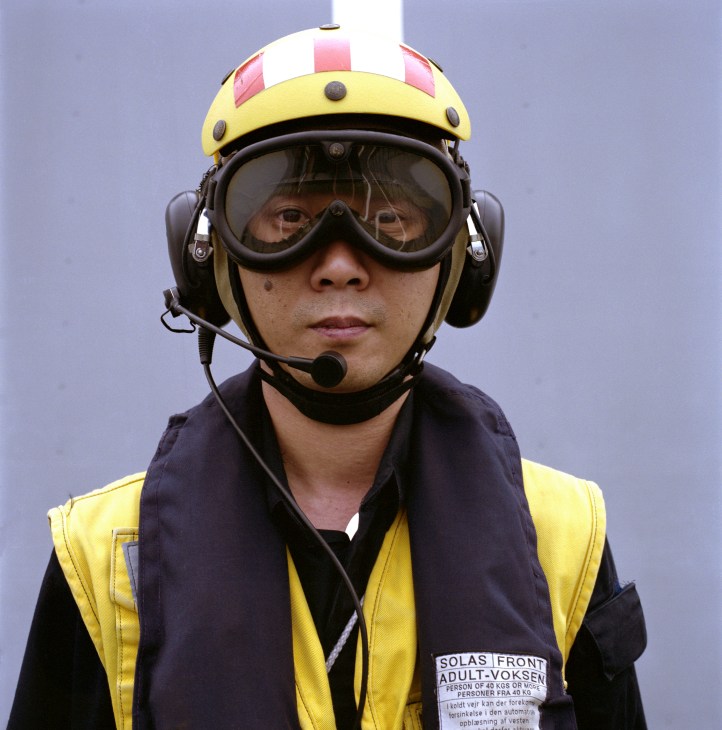 A serviceman wearing a yellow helmet with headset and goggles assists with helicopter landing operations.