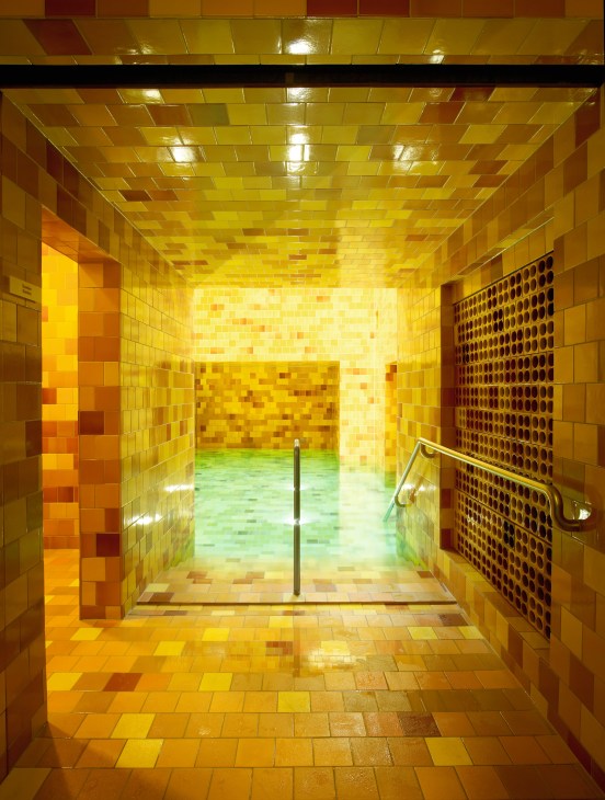Interior of Samedan spa showing yellow and gold tile-covered steam room with monochromatic mosaic patterns and green pool visible beyond.