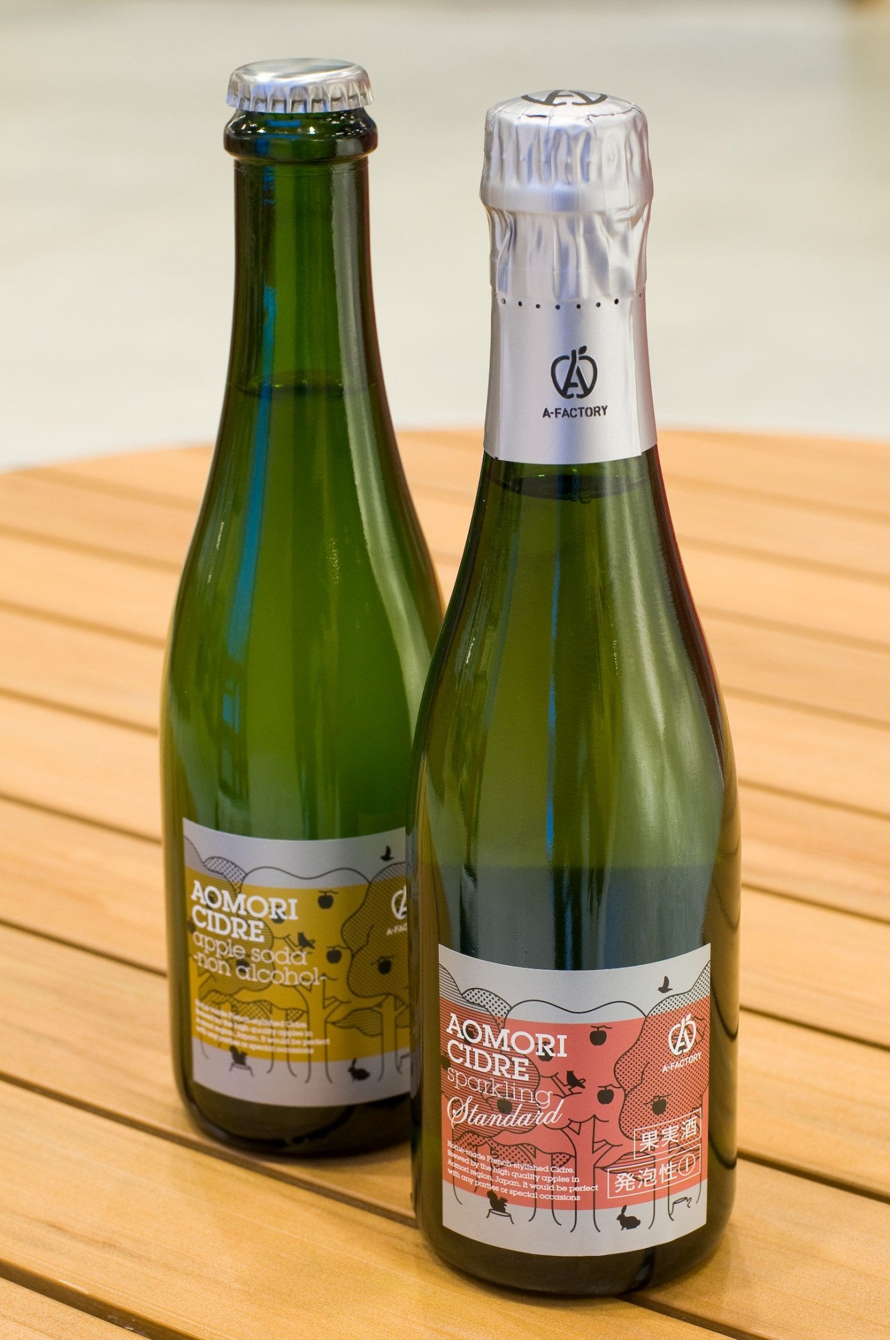 Two bottles of A-Factory Aomori Cidre with yellow and red labeled varieties on a wooden surface.