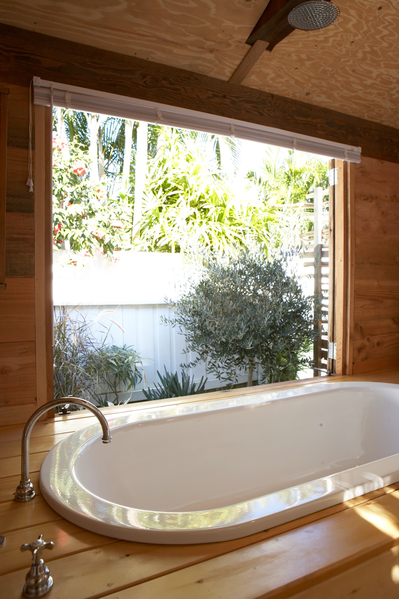 Wooden bathroom with oval bathtub and large window overlooking garden plants and water view.