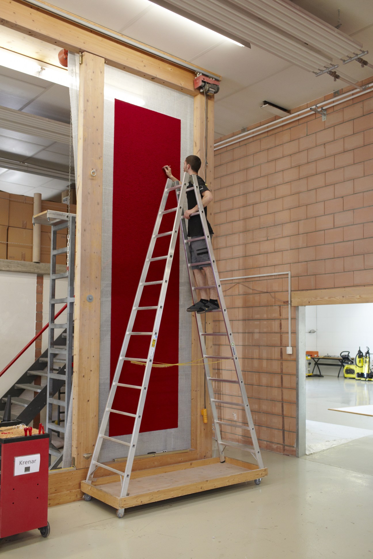 Worker on ladder adjusting a large wooden frame holding red carpet material in workshop.