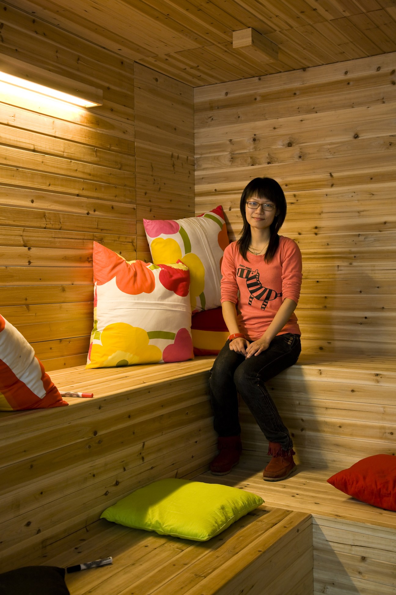 Young woman sitting in a wooden sauna-style room with colorful cushions.