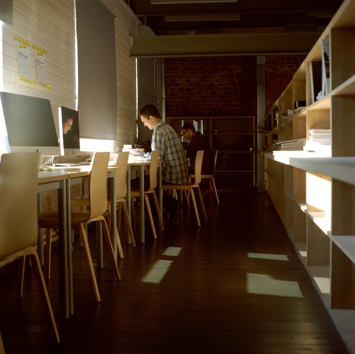 Interior view of Strelka Institute library with students working at wooden desks and illuminated shelving.