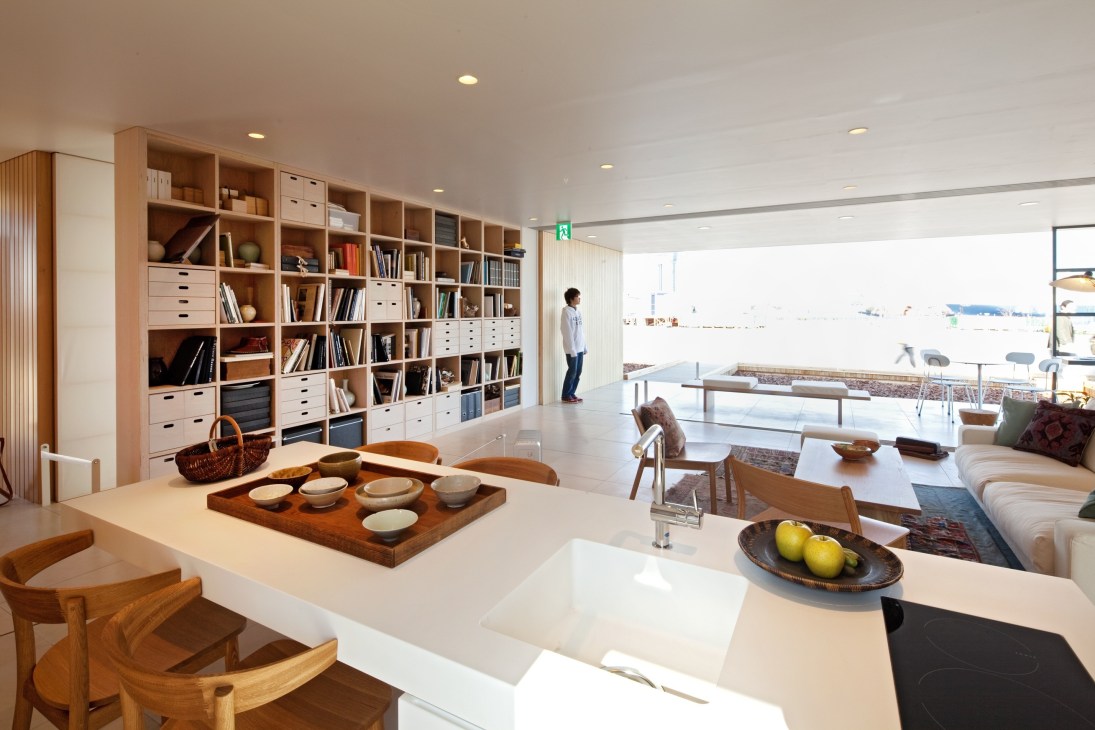 Modern minimalist living space with wooden shelving units serving as structural columns, white surfaces, and natural light from large windows.