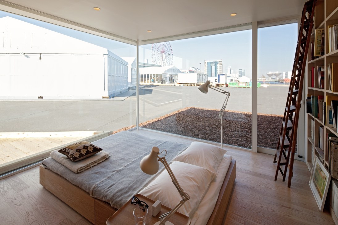 Minimalist bedroom with storage bed, desk lamp, and floor-to-ceiling windows overlooking Tokyo cityscape.