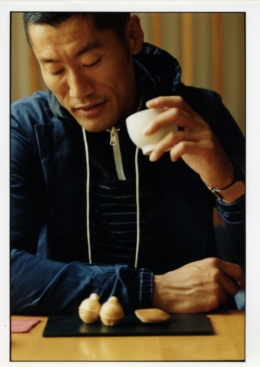 Model Yusuke wearing a Bottega Veneta jacket and Sacai hooded top, holding food at a table.