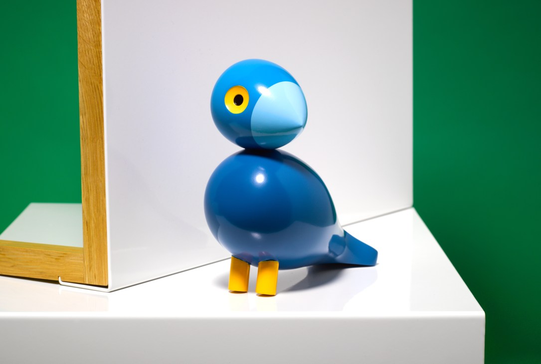 Blue wooden songbird toy with yellow eyes and feet by Kay Bojesen, displayed against a white and green backdrop.