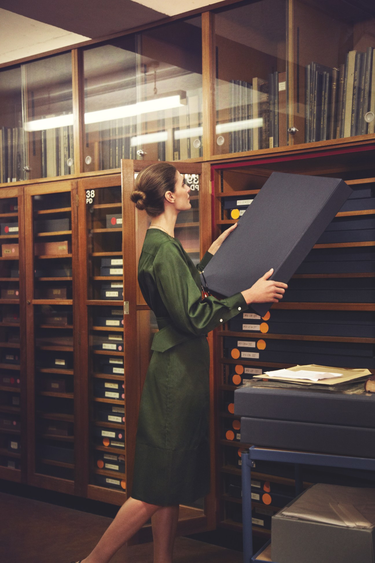 Woman in green Céline dress viewing microfiche in a library archive surrounded by wooden filing cabinets.