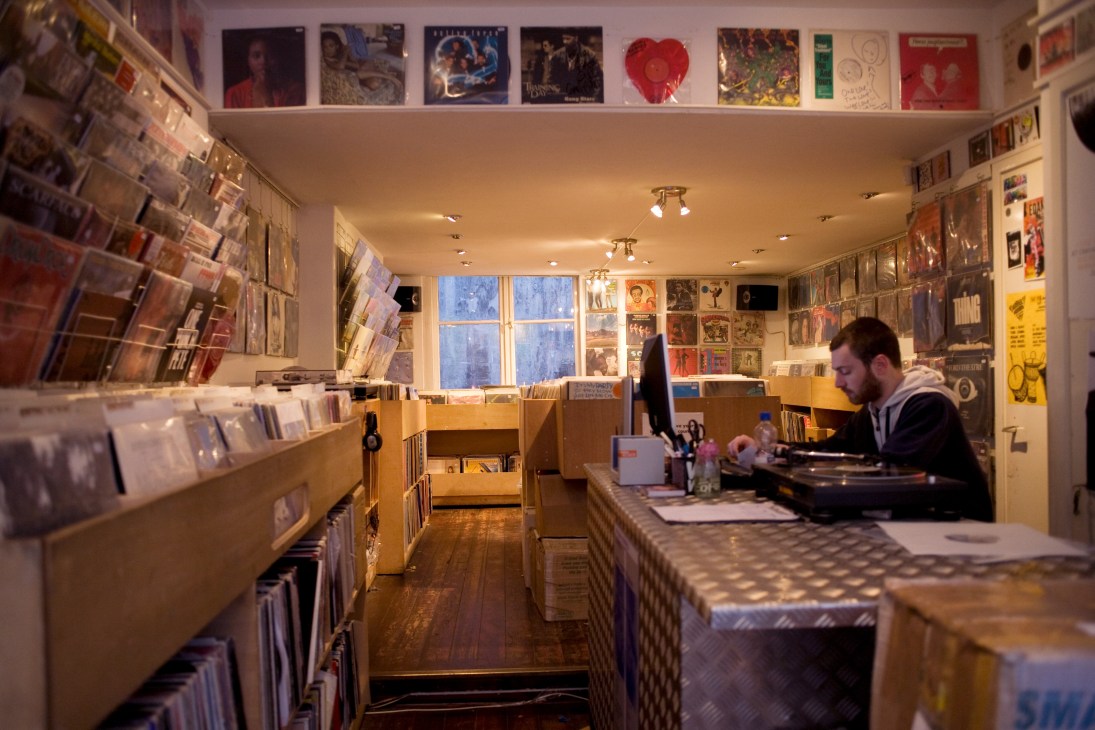 Interior of Waxwell Records in Amsterdam showing vinyl records on wall displays and a male staff member behind the counter.