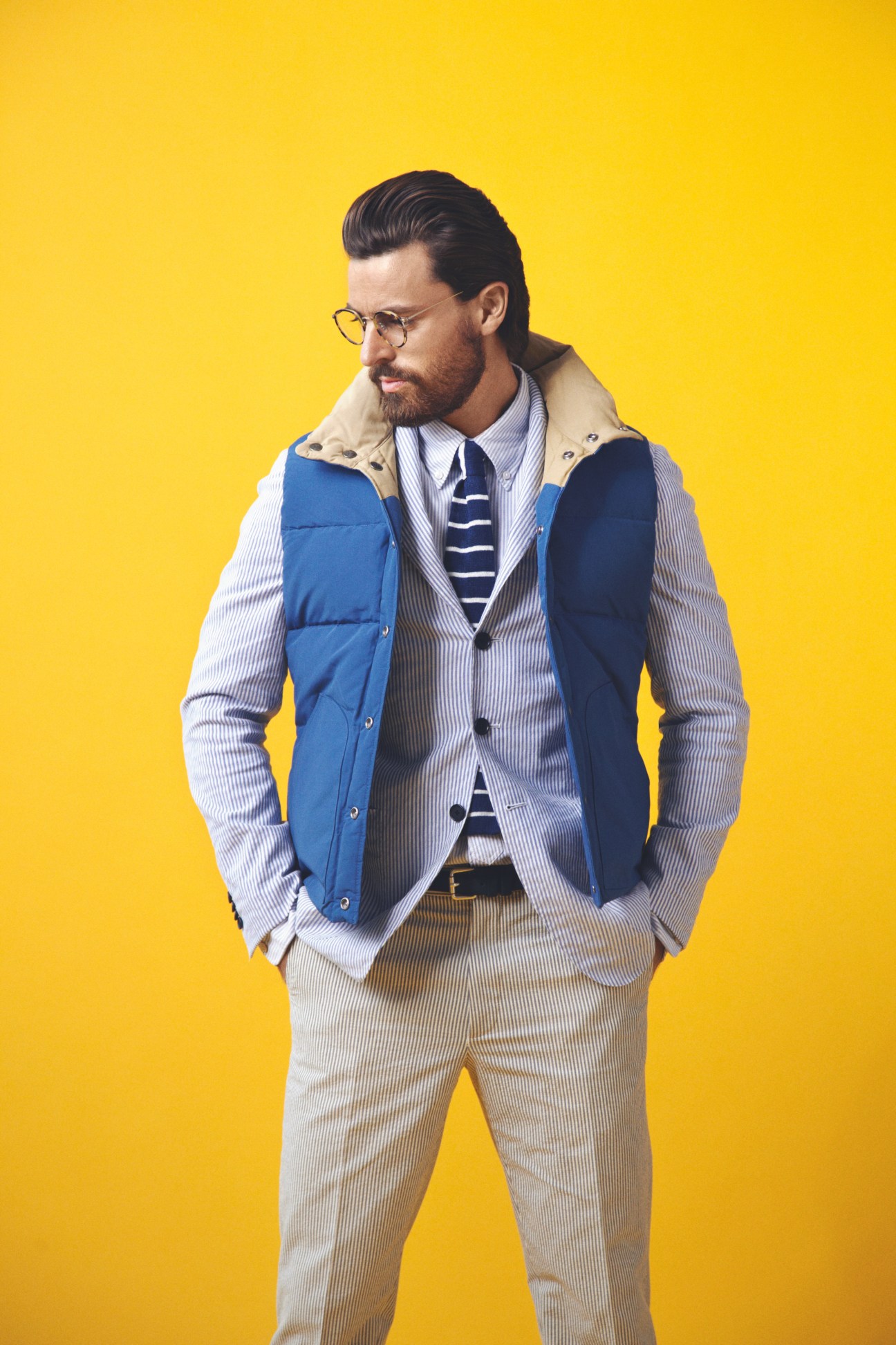 Male model wearing Oliver Peoples glasses, Mt Rainier Design vest, Caruso jacket, and Engineered Garments shirt against yellow background.