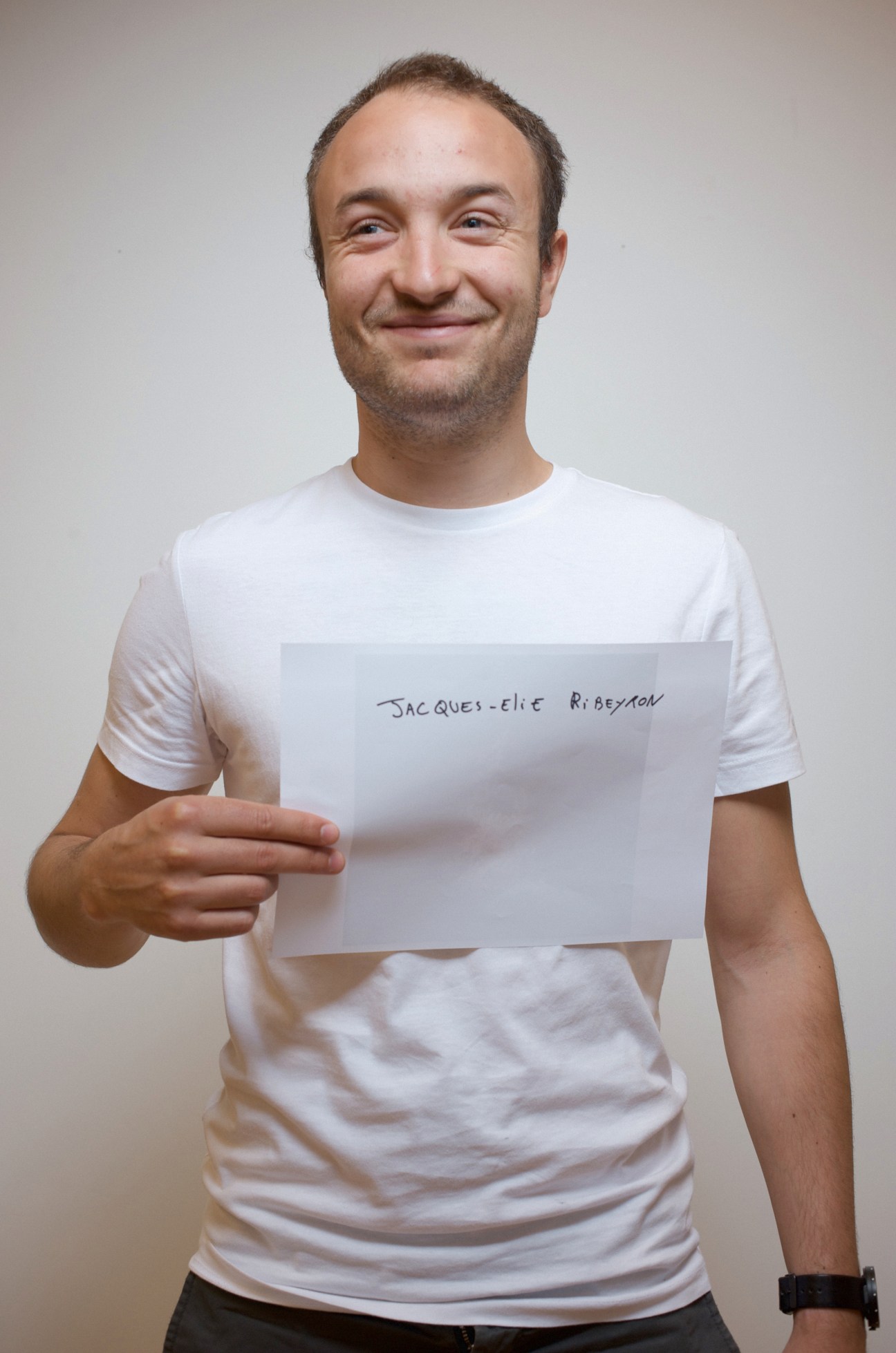 Man in white t-shirt holding a handwritten name card identifying him as Jacques-Elie Ribeyron.