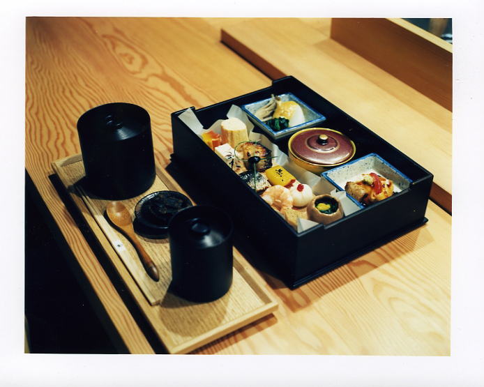 A Japanese bento box with multiple compartments containing tofu and vegetables on a wooden table with miso soup and chopsticks.