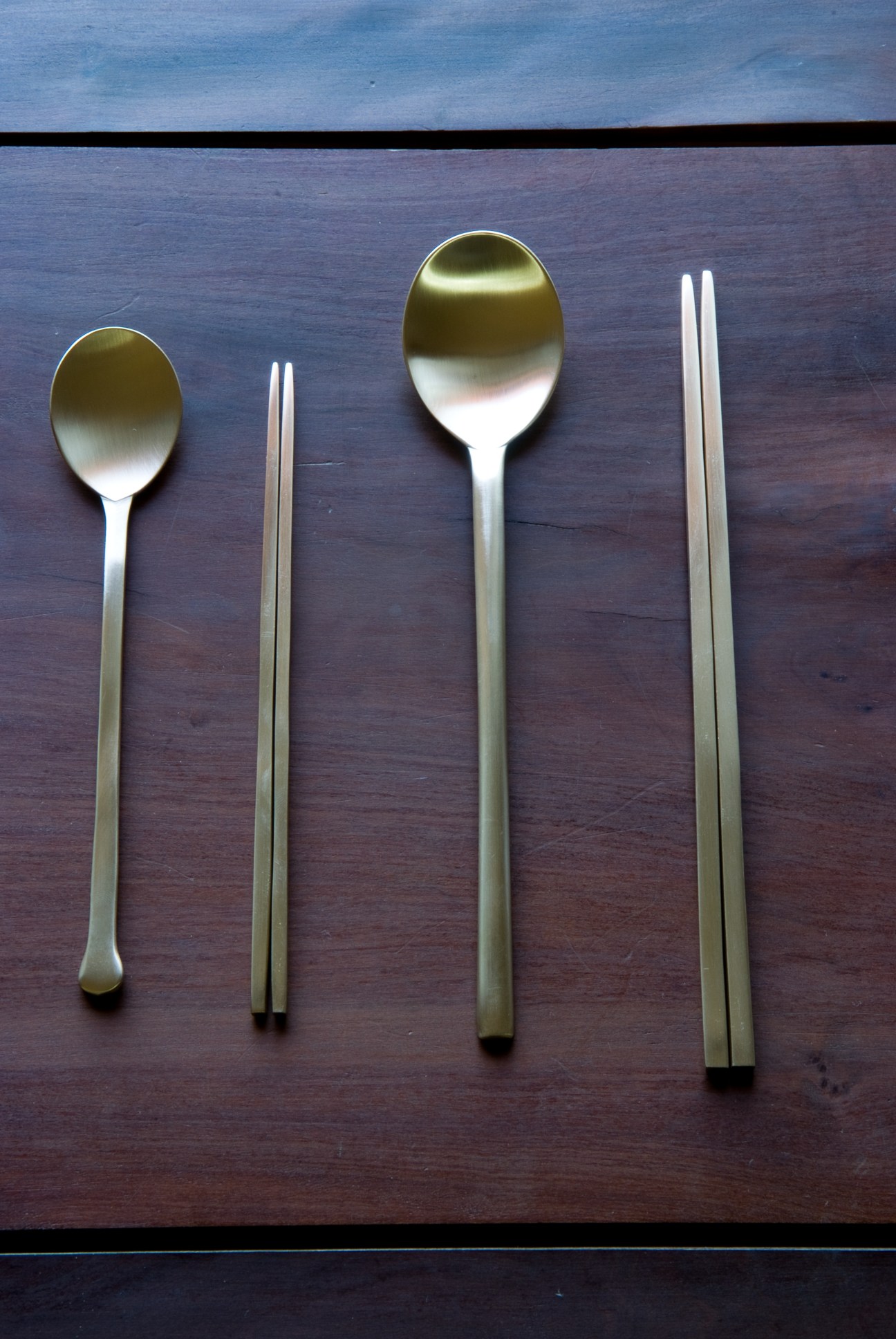 Two spoons and two pairs of flat metal chopsticks arranged on a dark wooden surface with white-gold finish.