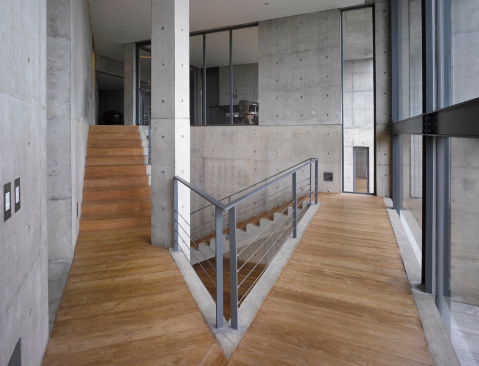 Interior hallway with wooden flooring, concrete walls, metal railing, and angular architectural elements.