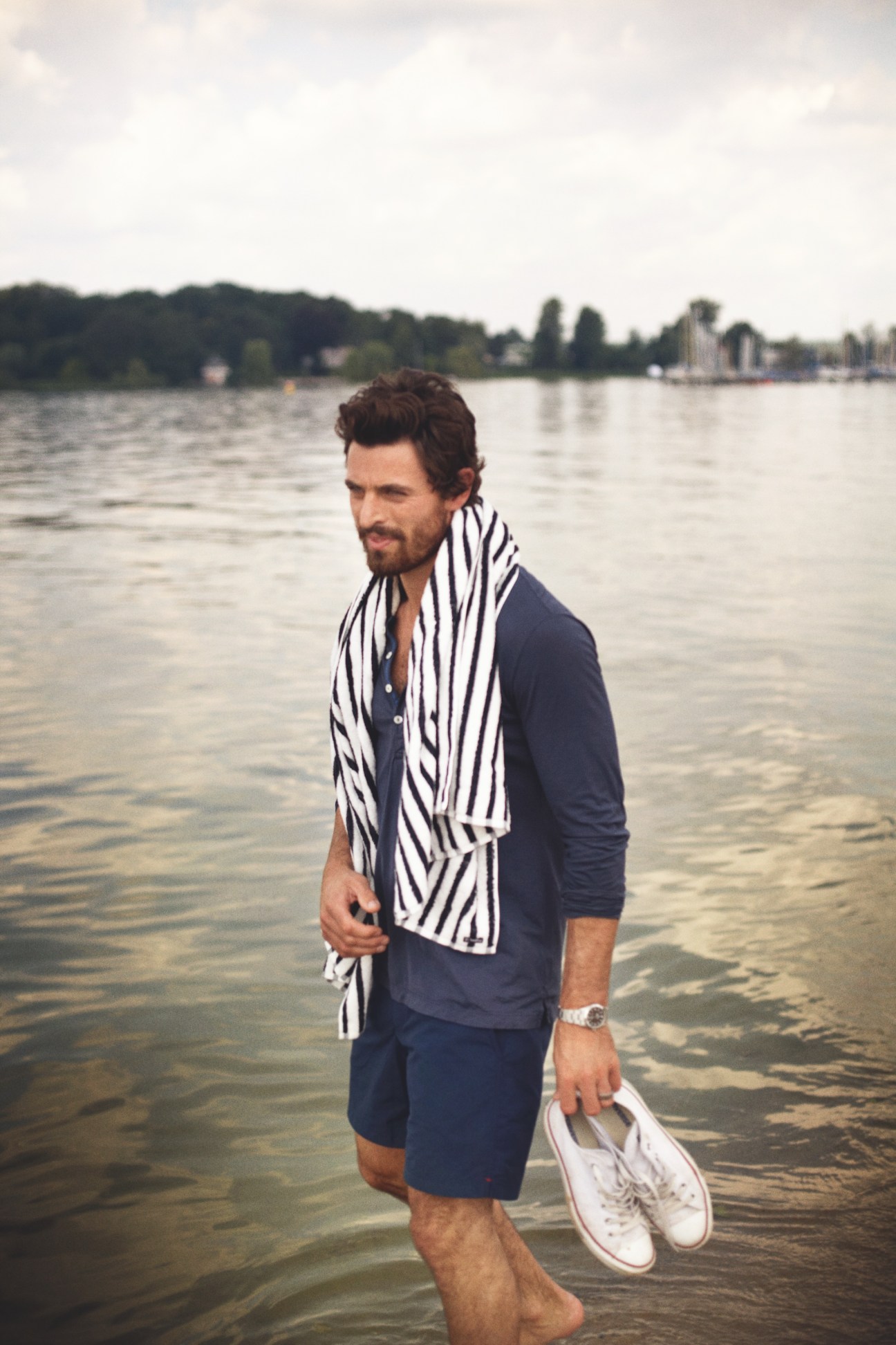Man wading in lake water wearing striped scarf, navy shirt and shorts, holding white sneakers.